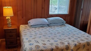 Een slaapkamer met houten lambrisering, een bed met een bloemendecoreerde spreideken, een nachtkastje met een lamp en een raam met jaloezieën.