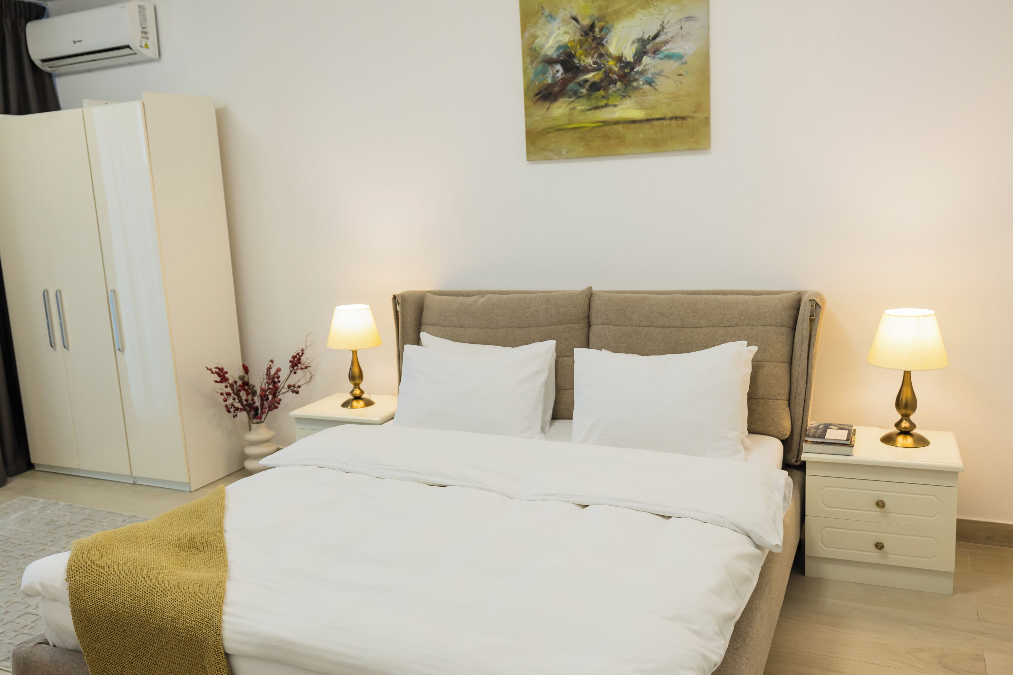 Un lit bien fait, avec du linge de lit blanc, deux lampes de chevet, une table de chevet blanche, une tête de lit beige, une armoire blanche et un tableau accroché au mur.