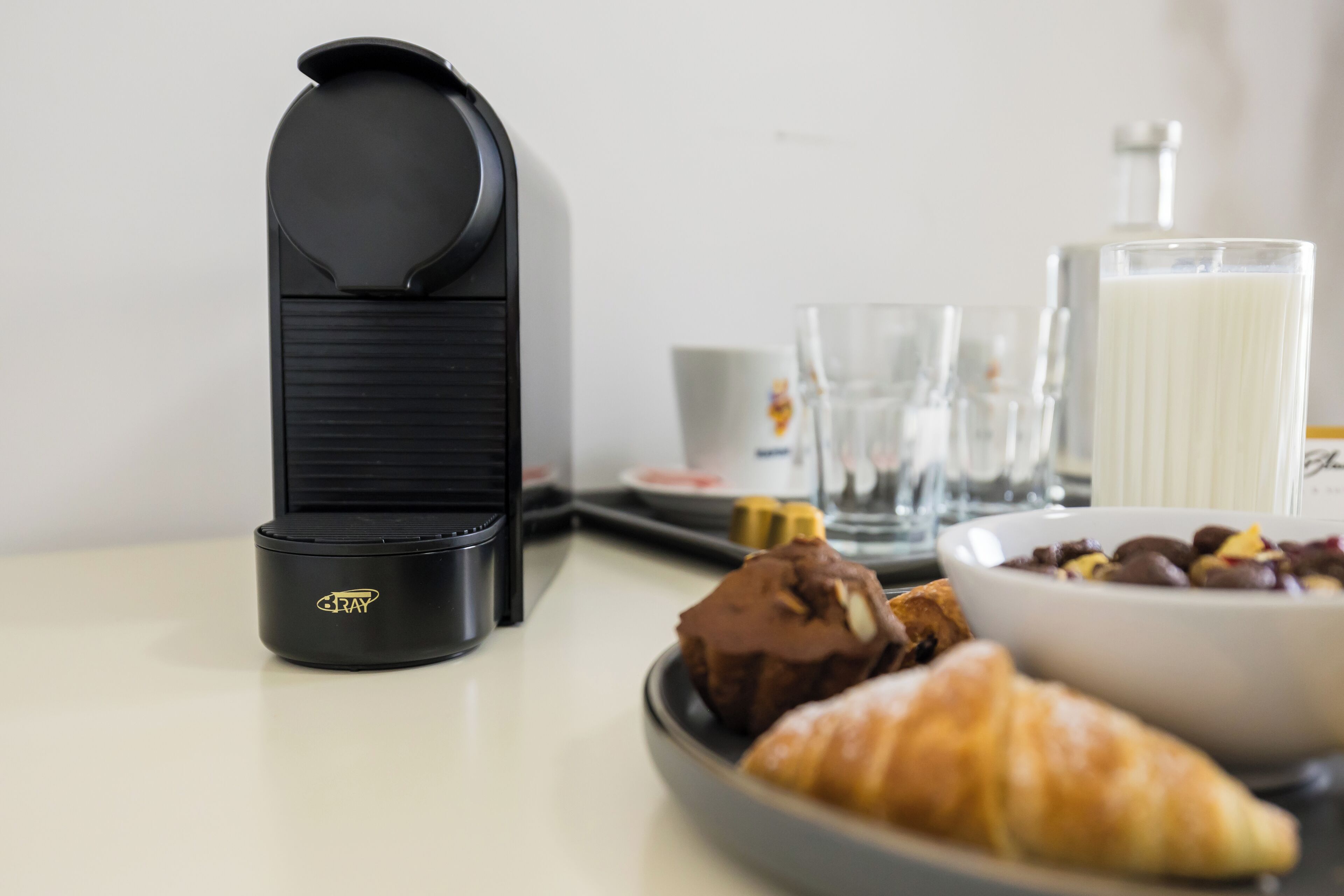 Una escena de desayuno con una cafetera, croissants, un tazón de chocolate y un vaso de leche sobre una mesa.