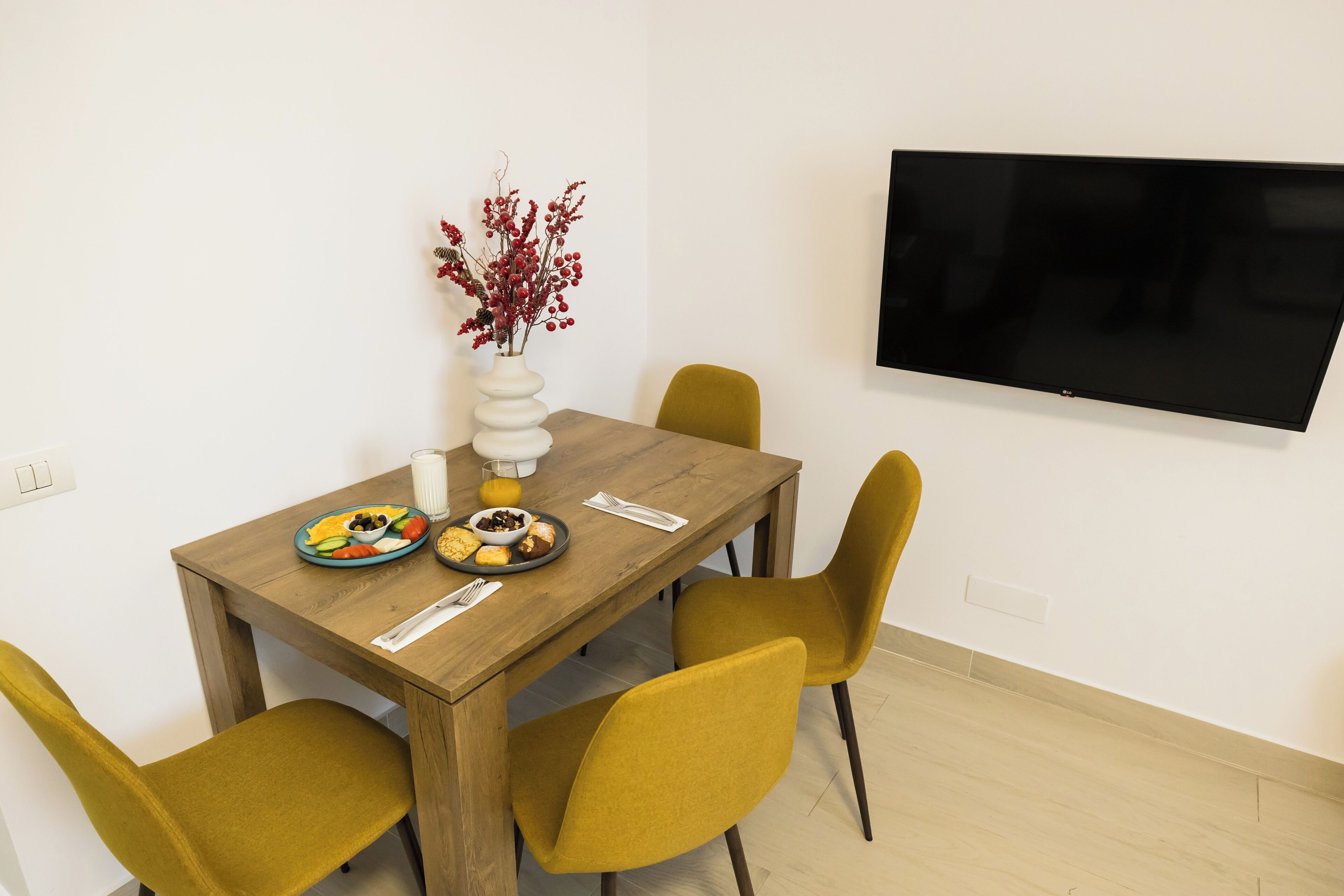 A dining area with a wooden table, four mustard-colored chairs, a vase with red berries, and a flat-screen TV mounted on the wall.