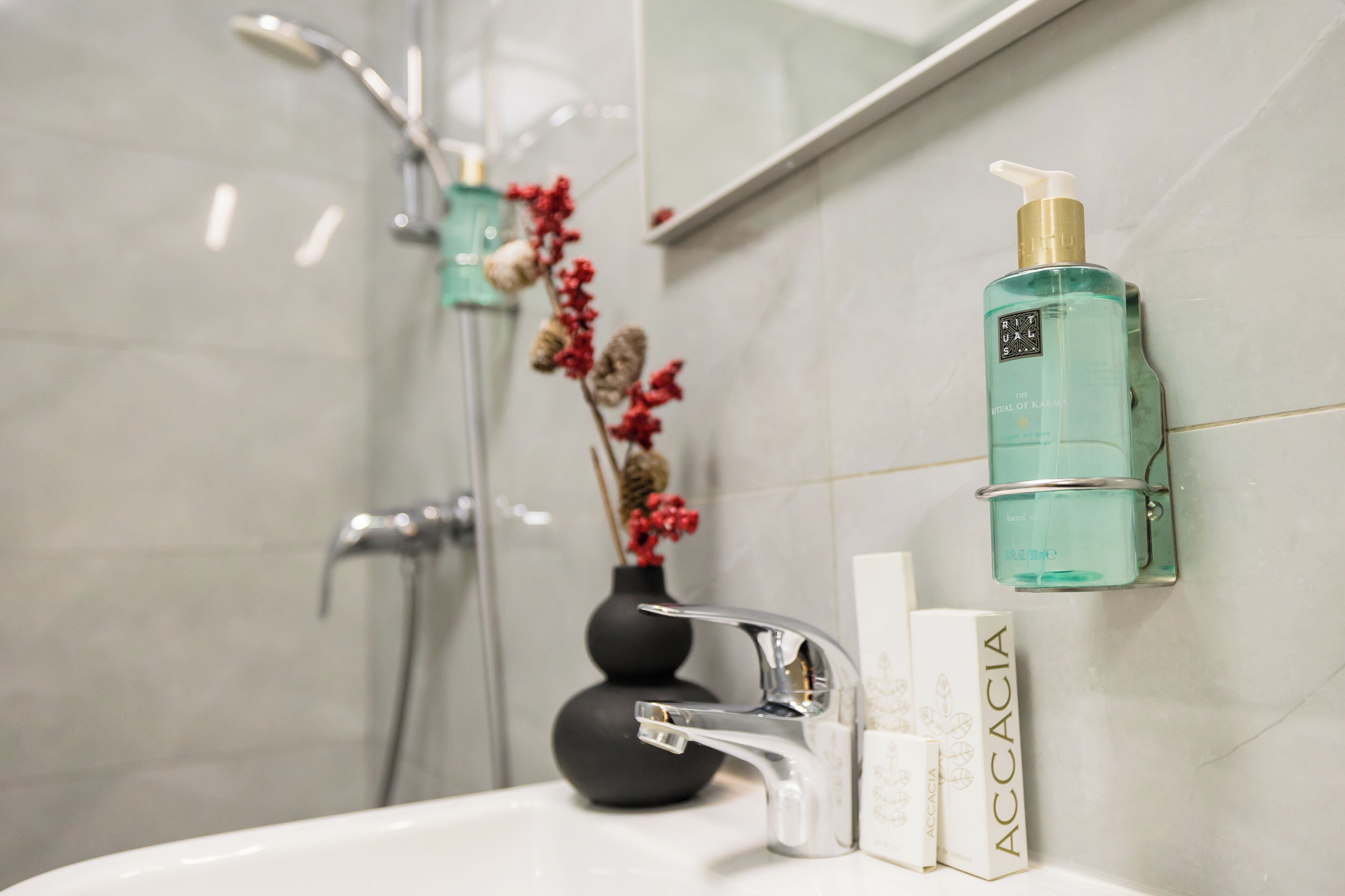 A bathroom with a white sink, a silver faucet, a green bottle of Accacia body wash, and a black vase with red flowers.
