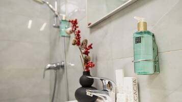 A bathroom with a white sink, a silver faucet, a green bottle of Accacia body wash, and a black vase with red flowers.