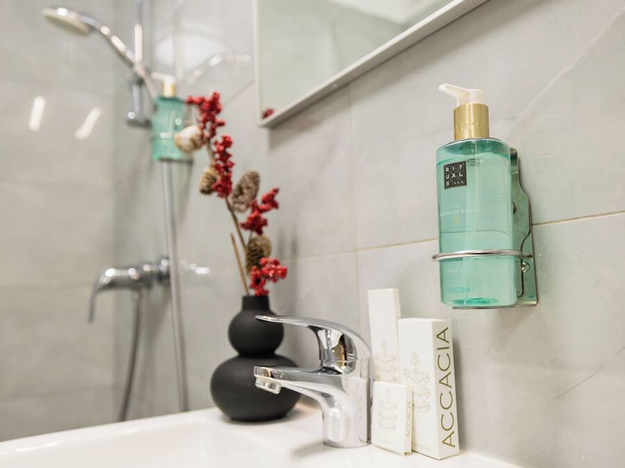 A bathroom with a white sink, a silver faucet, a green bottle of Accacia body wash, and a black vase with red flowers.