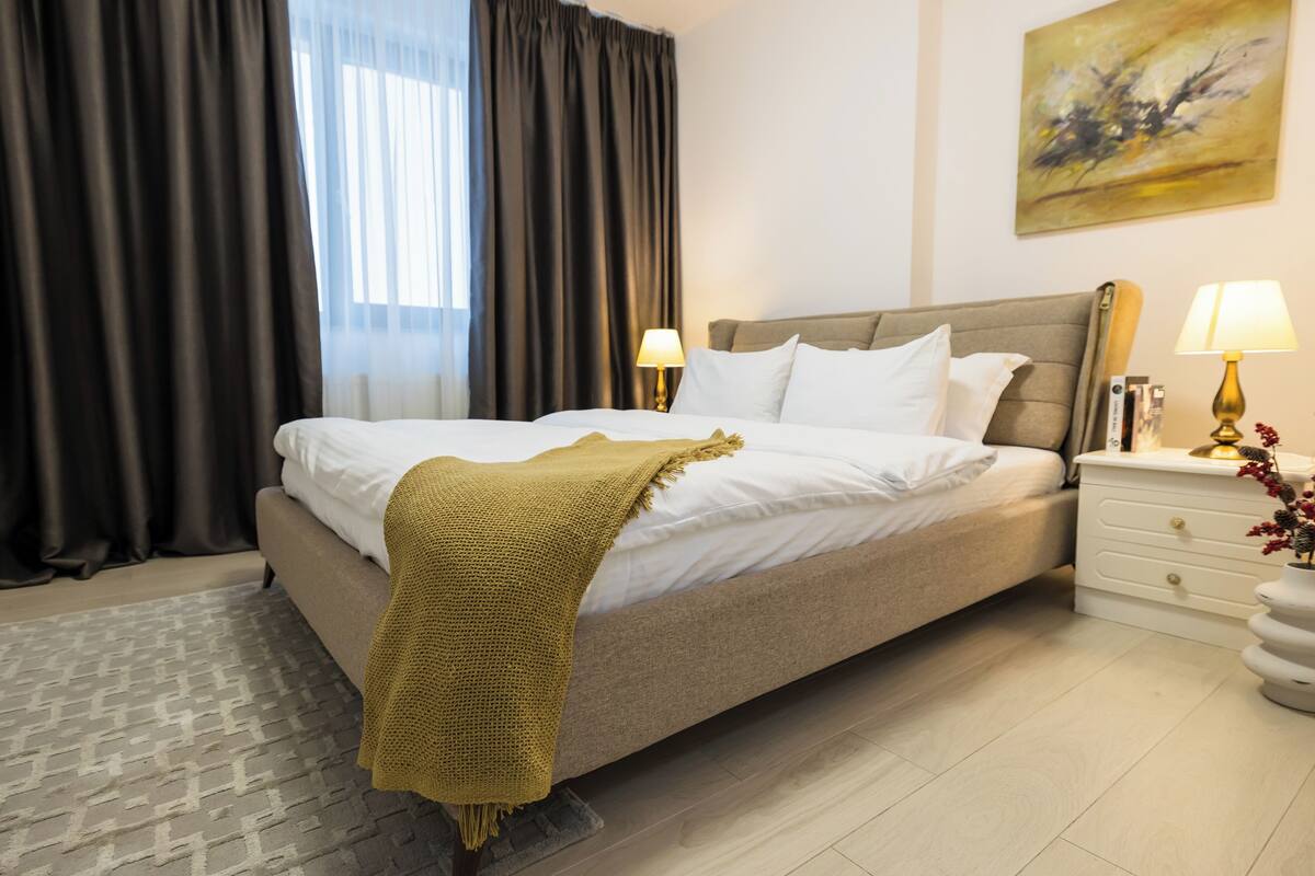 A neatly made bed with white linens, a beige headboard, and a mustard-colored throw blanket. There are two bedside tables with lamps and a small vase with flowers. The room has a patterned rug and a painting on the wall.