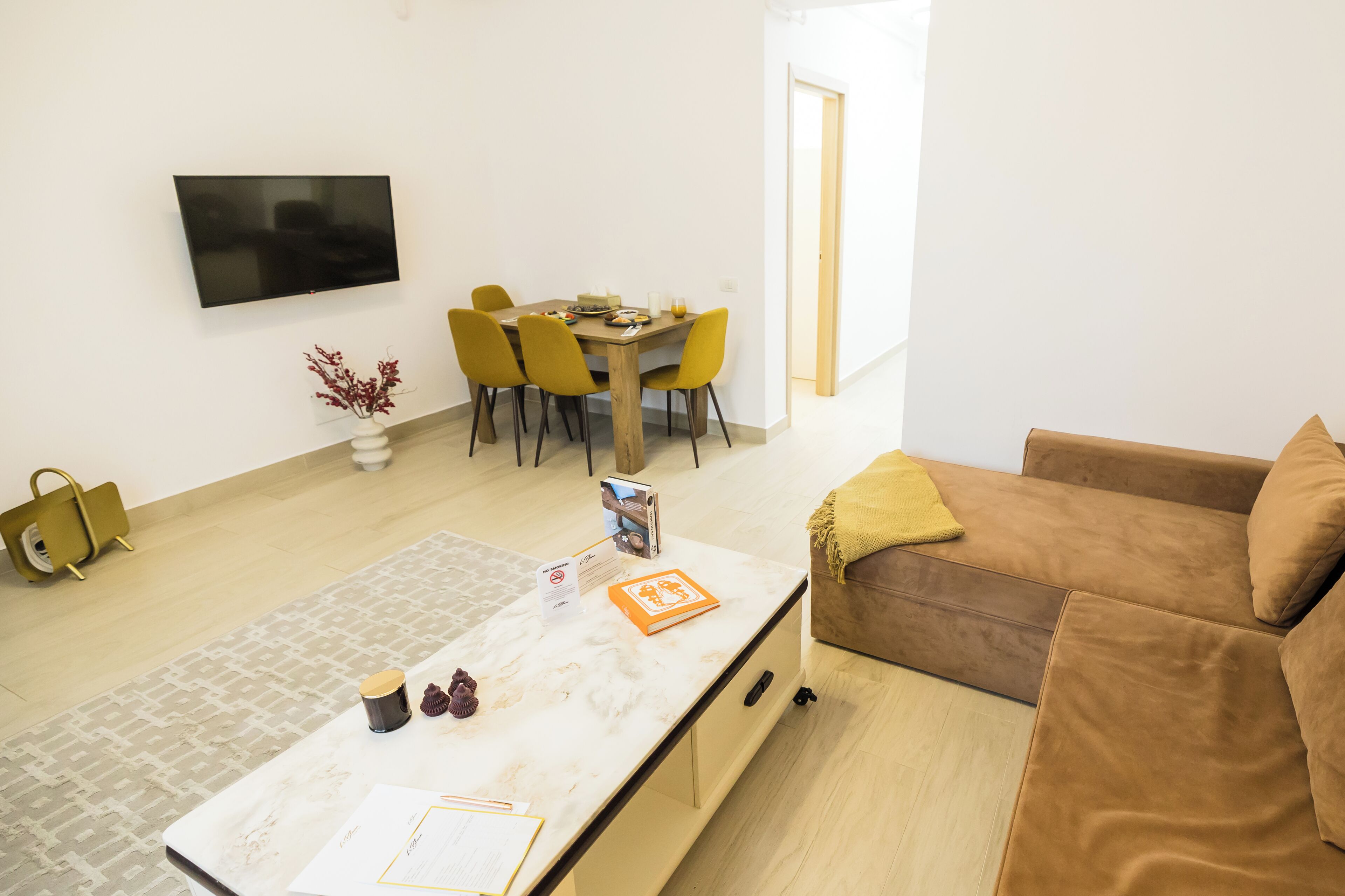 A modern living room with a sofa, a dining table with chairs, a TV mounted on the wall, and a small plant.