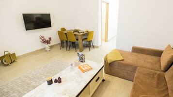 A modern living room with a sofa, a dining table with chairs, a TV mounted on the wall, and a small plant.