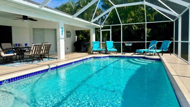Una piscina azul cristalina con una zona de terraza con techo blanco, sillas de descanso y un ventilador de techo.