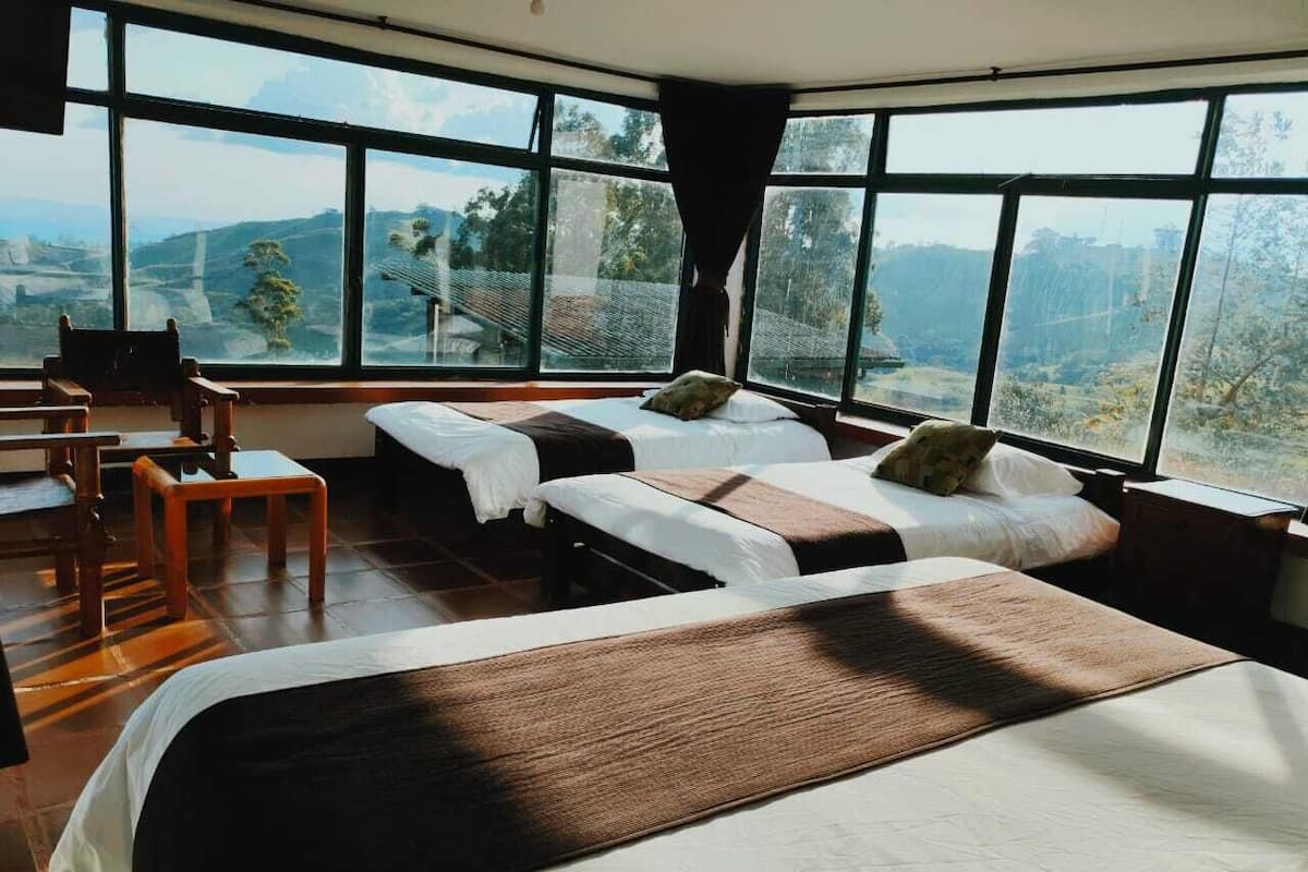 A hotel room with two beds, a small table, and a chair. The room has large windows offering a view of the outdoors.
