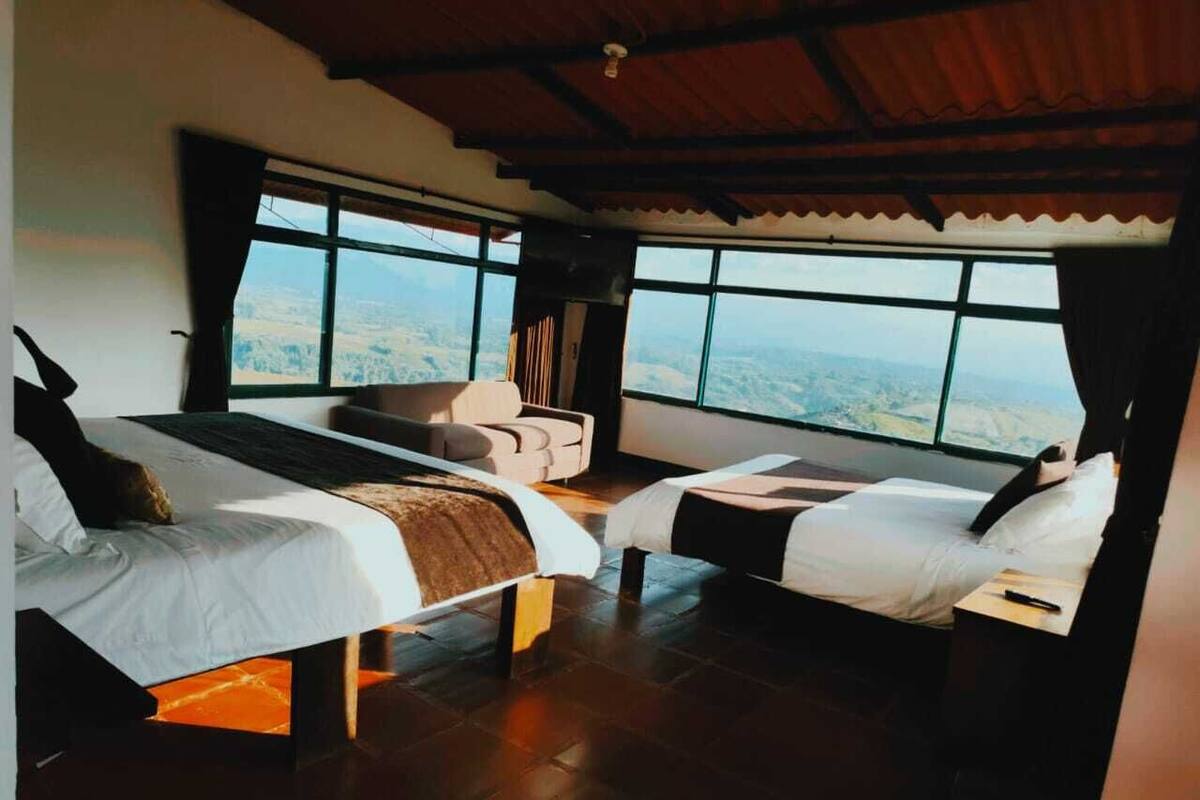 A room with two beds, a sofa, and a large window offering a view of the landscape.