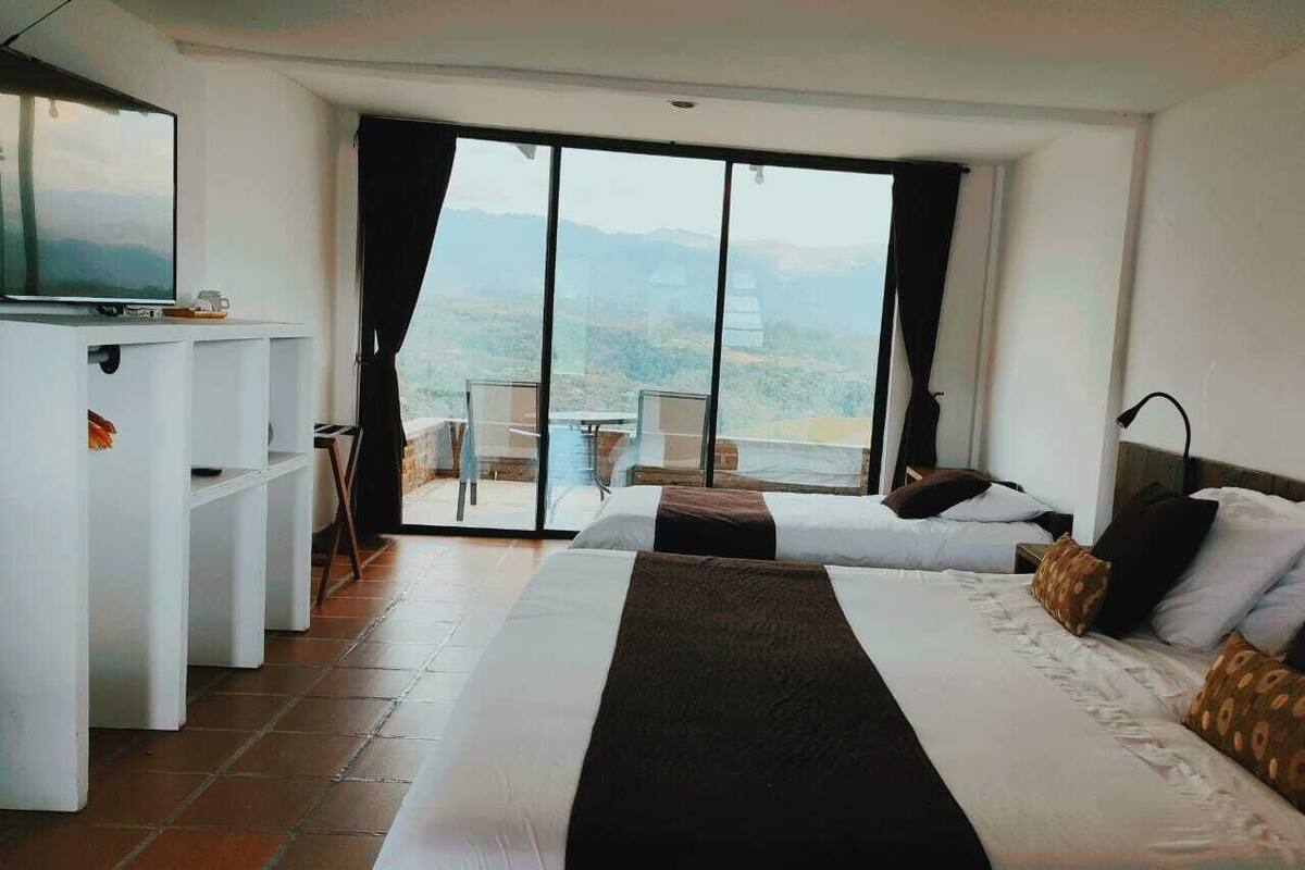 A hotel room with a large bed, a TV, a desk, and a balcony with a view of the mountains.