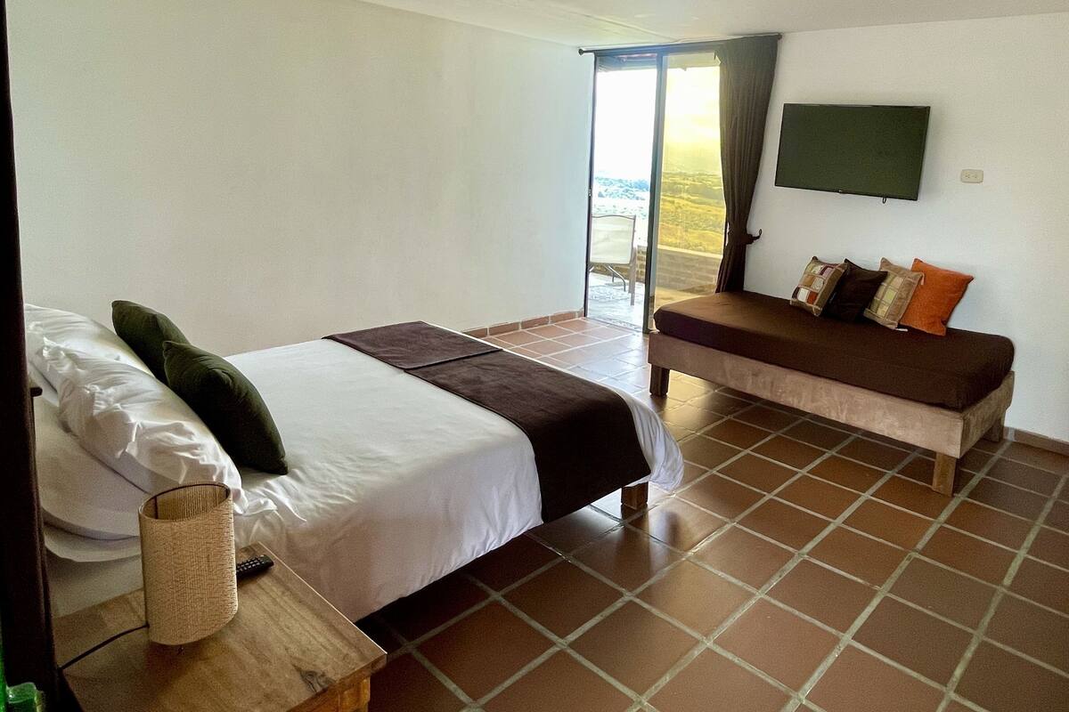A bedroom with a bed, a sofa, a TV, and a view through a glass door.