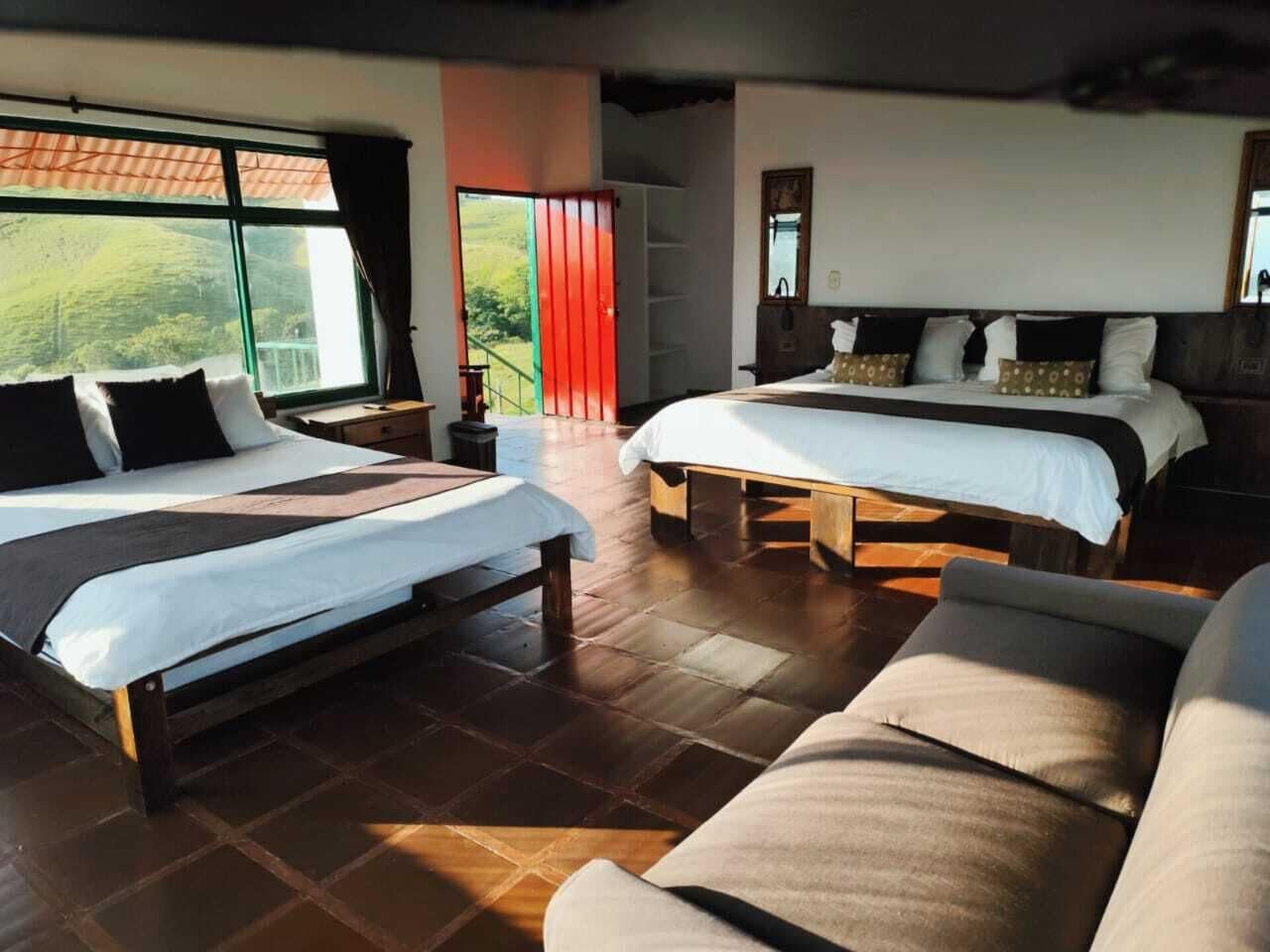 Camera d'albergo con due letti, un divano e un'ampia finestra che si affaccia su spazi verdi.