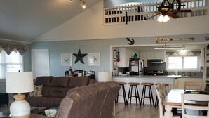 Ein großzügiges Wohnzimmer mit einer braunen Ledersofa, einem Holztisch und einem Essbereich mit weißem Tisch und Stühlen. Im Hintergrund ist eine Küche mit modernen Geräten zu sehen.