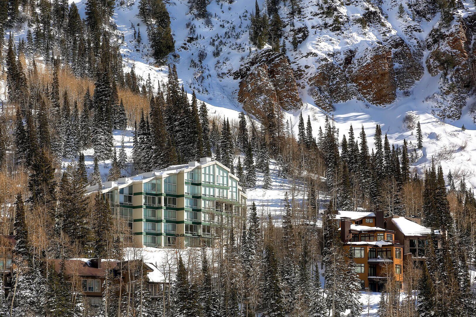 A modern hotel with a glass facade, surrounded by snow-covered mountains and pine trees.