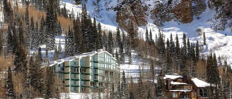 A modern hotel with a glass facade, surrounded by snow-covered mountains and pine trees.