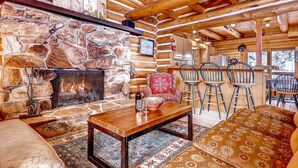 Ein Blockhaus-Interieur mit Kamin aus Stein, Holzbalkendecke, einer Essecke mit Barhocker und einer gemütlichen Sitzgruppe.