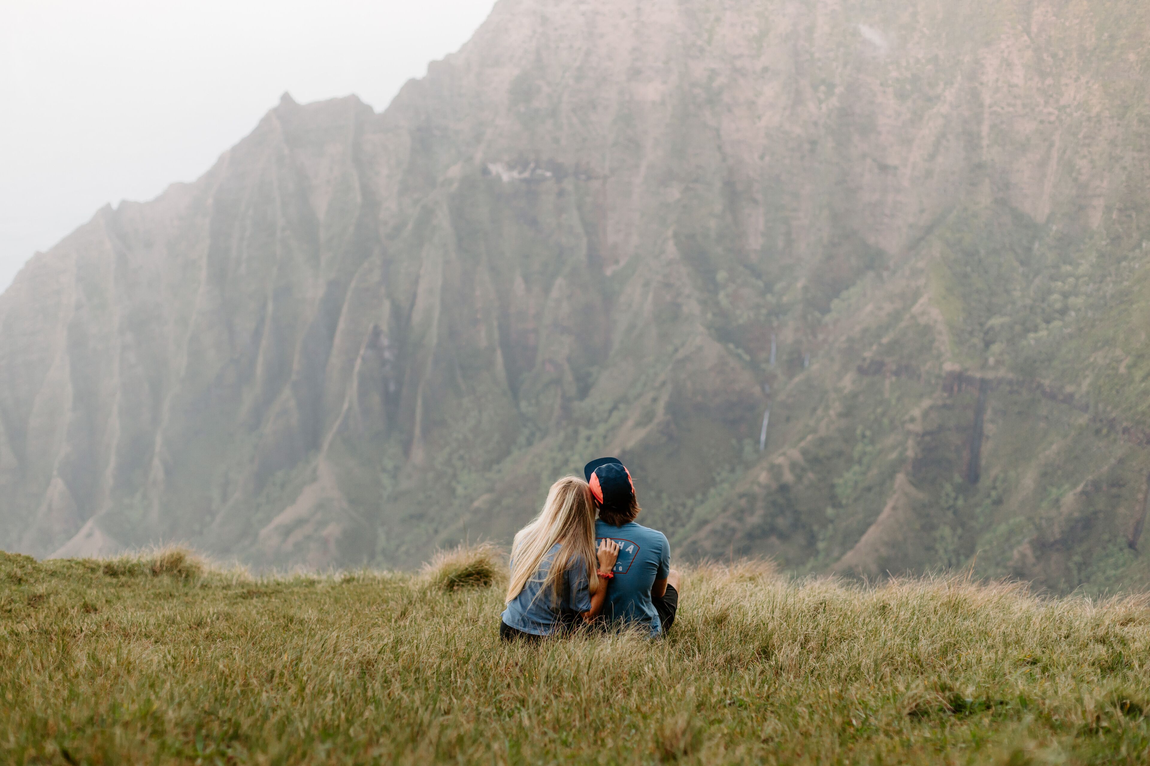 Un couple est assis sur une colline herbeuse, avec une chaîne de montagnes en arrière-plan.