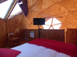 Una habitación acogedora con techo de madera inclinado, cama con ropa de cama roja y blanca, televisor montado en la pared y ventana que proporciona luz natural.