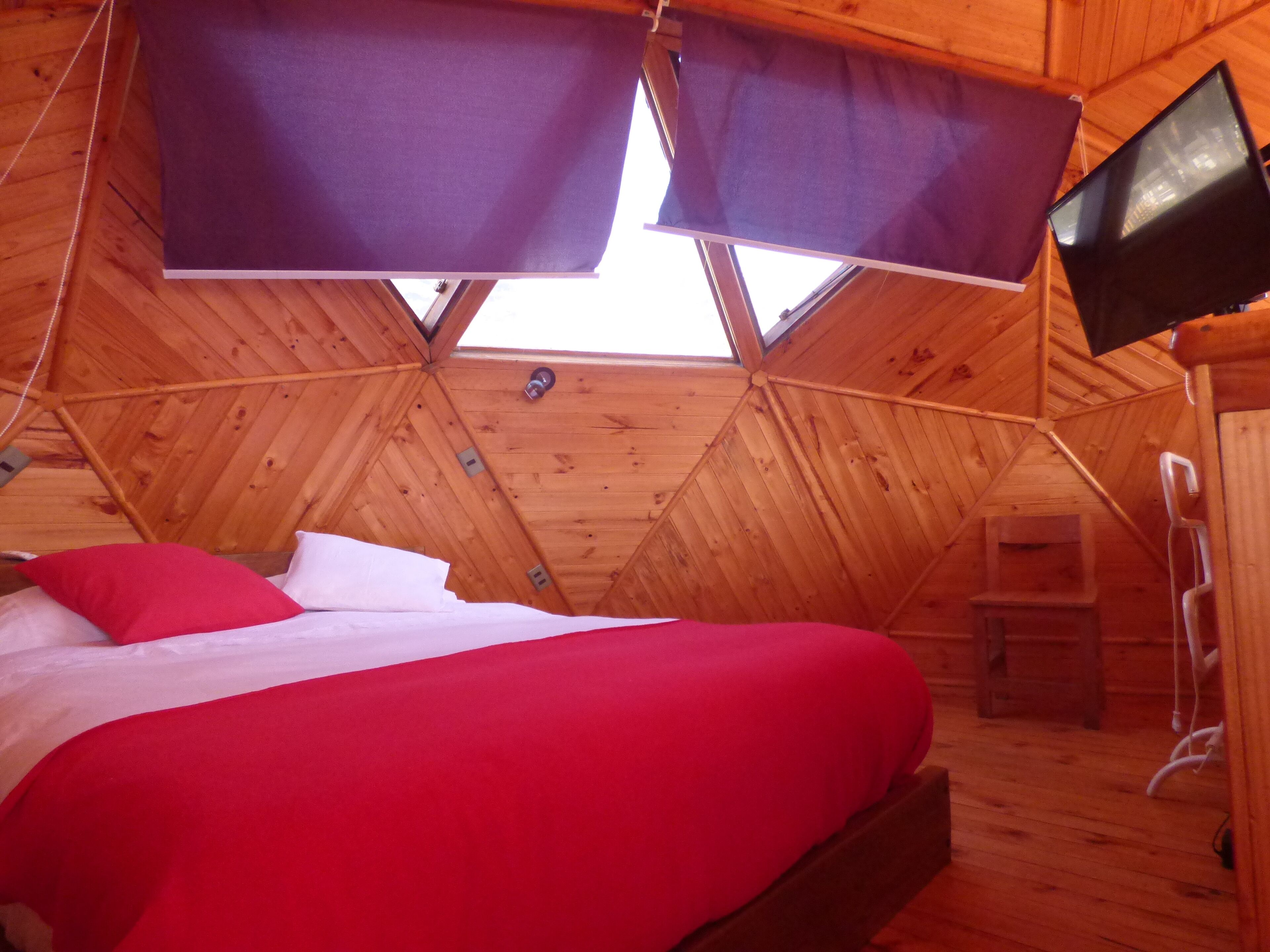 Acogedora habitación tipo cabaña de madera con una cama, un televisor montado en la pared y un tragaluz.