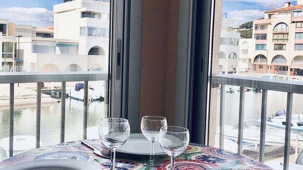 A balcony with a table set for two, overlooking a marina and buildings. - Charming, colourful and functional T2 apartment 200m from the beach free, enclosed parking (Sète)