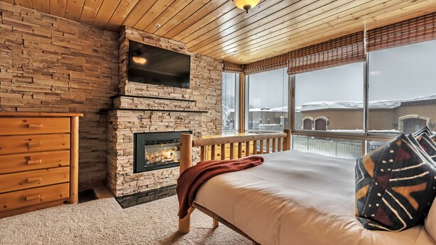 A bedroom with a stone fireplace, a wooden dresser, a bed with a patterned pillow, and a large window with a view of a snowy landscape. - Updated Condominium with Fireplace, Wine Fridge and Granite Countertops (Alta)