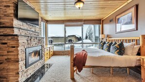 A cozy bedroom with a stone fireplace, a large window with a view of a snowy landscape, and a bed with patterned pillows. - Updated Condominium with Fireplace, Wine Fridge and Granite Countertops (Alta)
