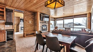 A cozy cabin interior with a stone fireplace, wooden dining area, and large windows offering a view of the snowy landscape. - Updated Condominium with Fireplace, Wine Fridge and Granite Countertops (Alta)