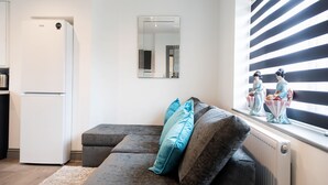 Una cocina moderna con un refrigerador blanco, un sofá gris con cojines turquesa, un espejo grande y un ventanal con persianas a rayas.