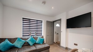 Una sala moderna con un sofá gris, cojines turquesa, un televisor de pantalla plana y una zona de comedor con mesa y sillas.