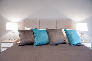 A neatly made bed with a grey quilt and four pillows in shades of blue and grey. There are two bedside tables with lamps and a wall-mounted light fixture.