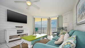 A living room with a teal sofa, a green coffee table, a flat-screen TV, and a ceiling fan. - 5th Floor 2 Bedroom with Reserved Phoenix 5 Parking (Orange Beach)