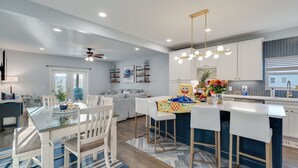 A modern kitchen with white cabinets, a central island with stools, and a dining area with a glass table and chairs. - Beach Side “Crabby Paddy", Luxury family retreat 6br 4+ bath w pool+hot tub (Panama City Beach)