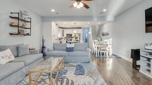 A modern living room with a sofa, a coffee table, and a ceiling fan. There is a kitchen in the background with a dining area. - Beach Side “Crabby Paddy", Luxury family retreat 6br 4+ bath w pool+hot tub (Panama City Beach)