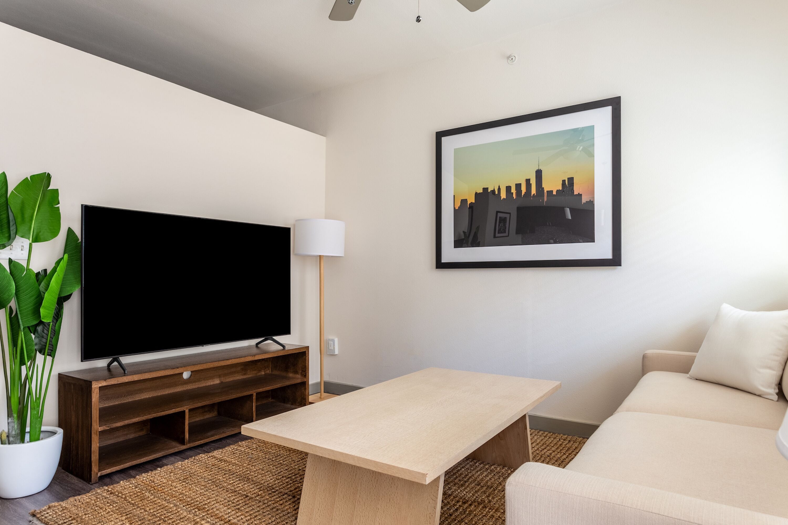 A modern living room with a flat-screen TV, a coffee table, a sofa, a framed cityscape picture, and a potted plant.
