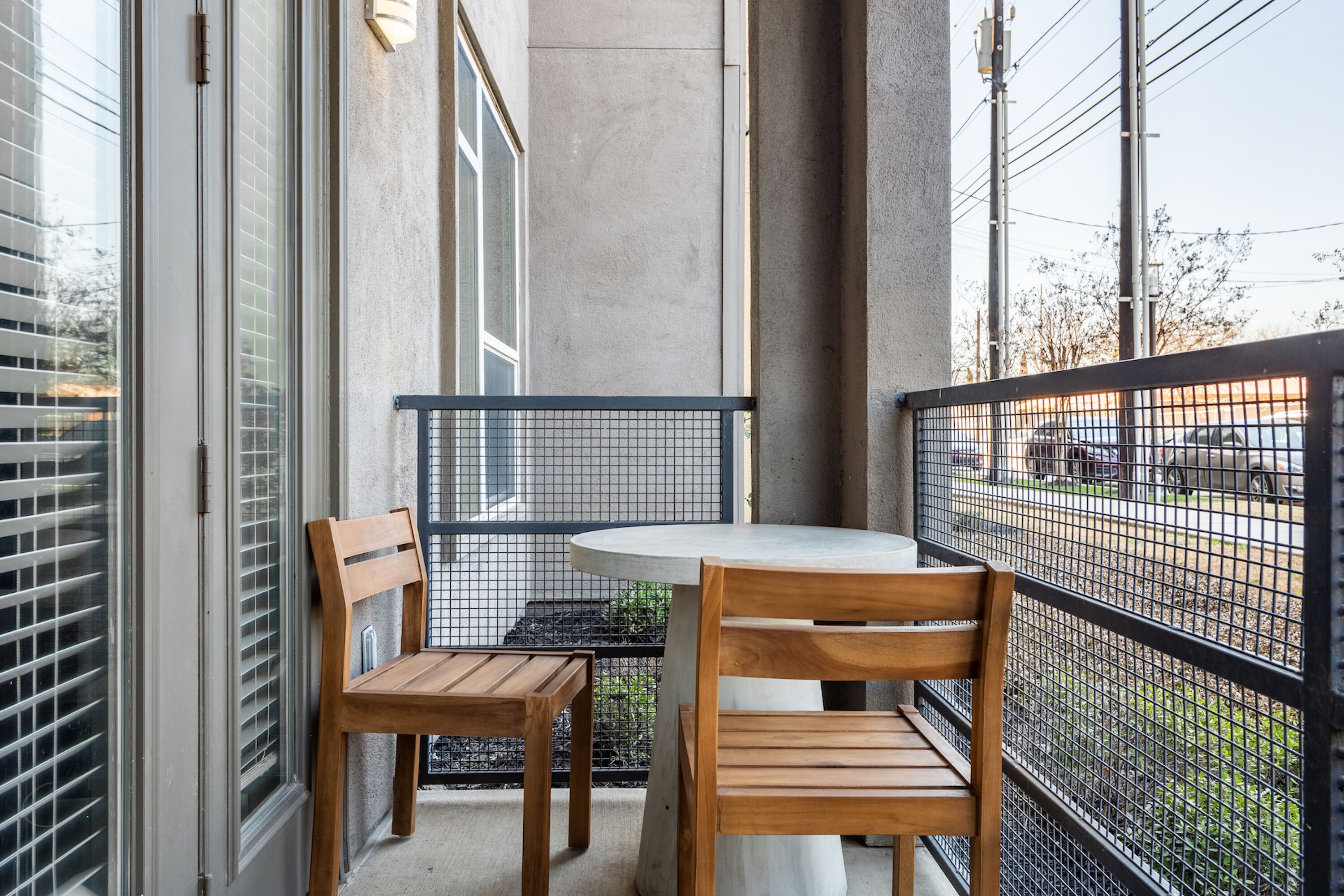 A balcony with a round table and two wooden chairs, a metal railing, and a view of the street and parked cars.