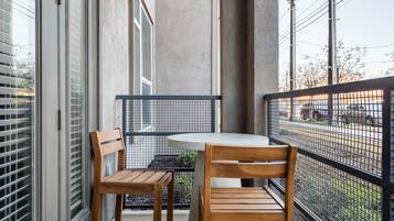 A balcony with a round table and two wooden chairs, a metal railing, and a view of the street and parked cars.