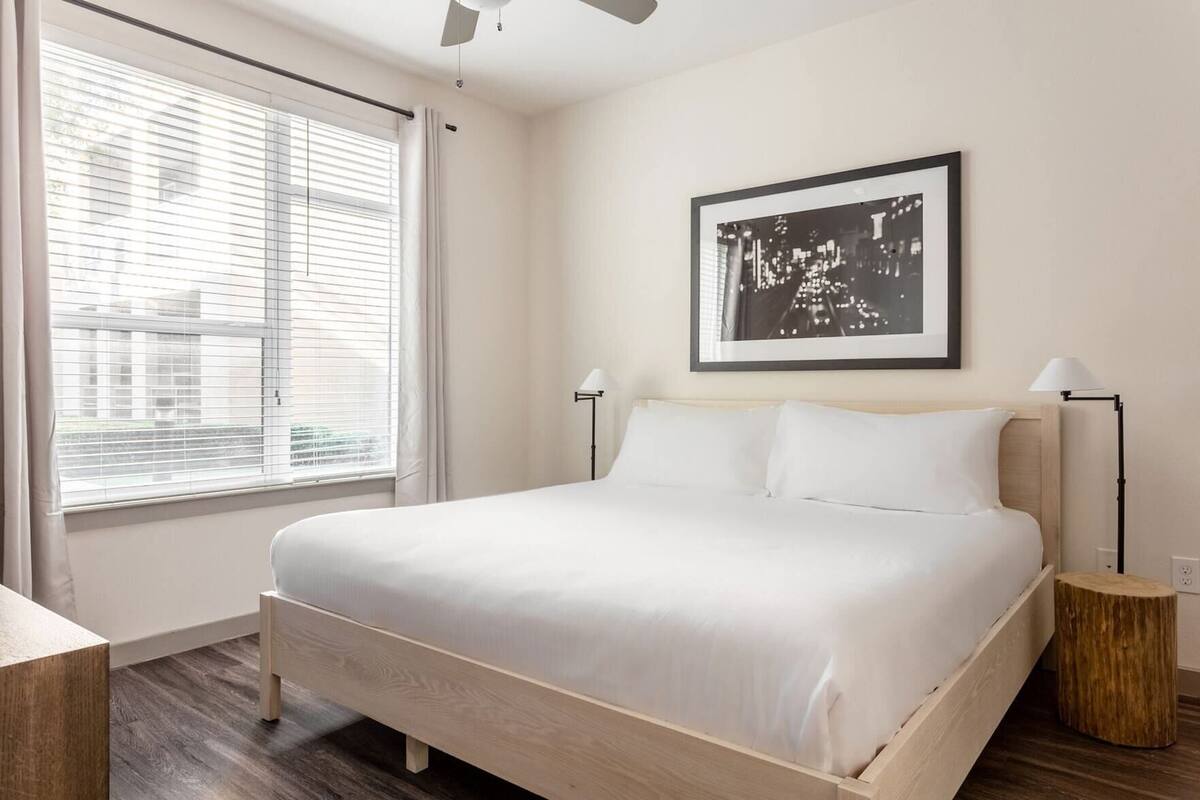 Une chambre à coucher avec un lit, une fenêtre avec des stores, un tableau encadré au mur et une lampe de chevet.