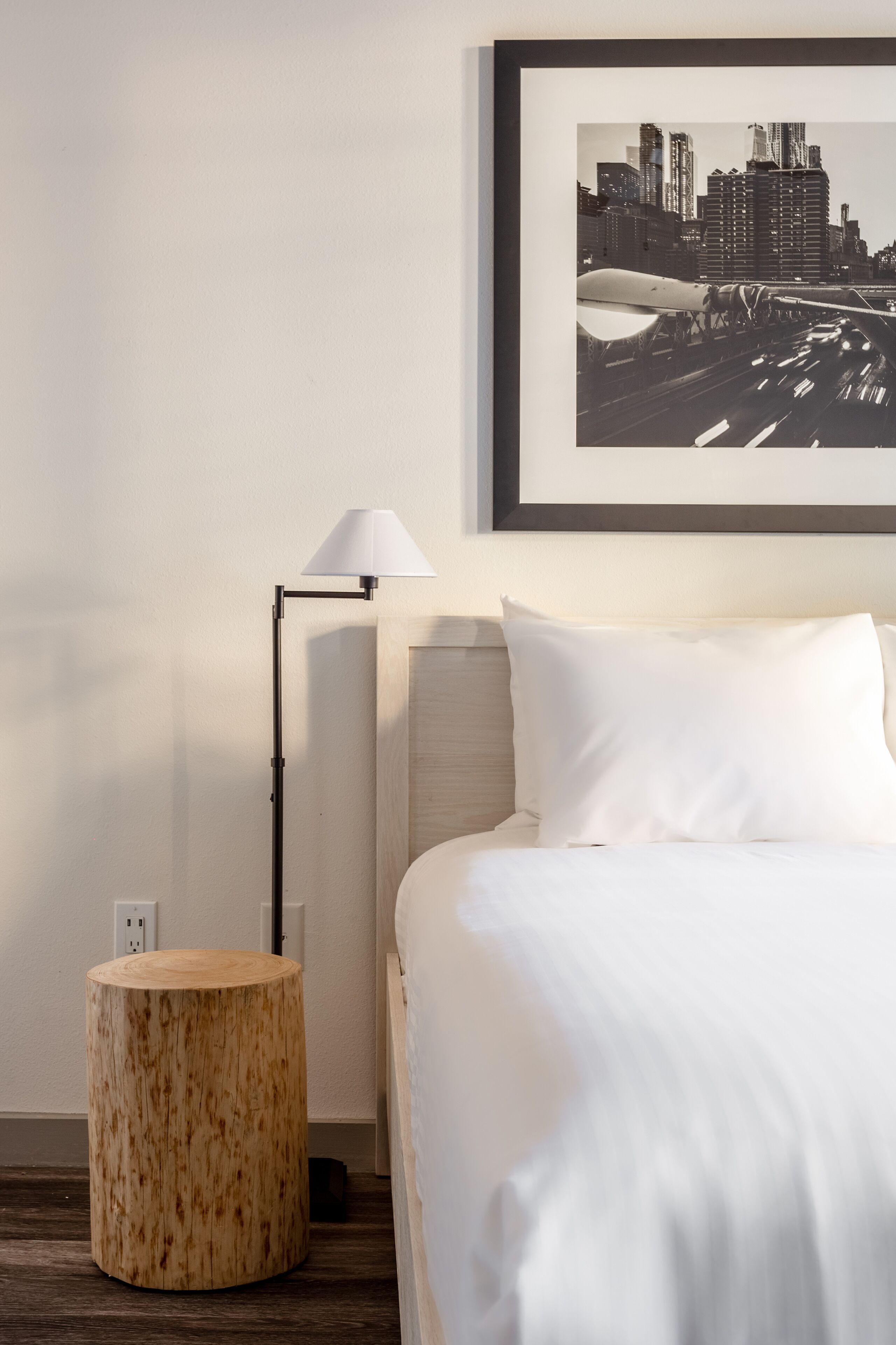 A hotel room with a bed, a round wooden side table, a floor lamp, and a framed cityscape picture on the wall.