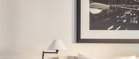 A hotel room with a bed, a round wooden side table, a floor lamp, and a framed cityscape picture on the wall.