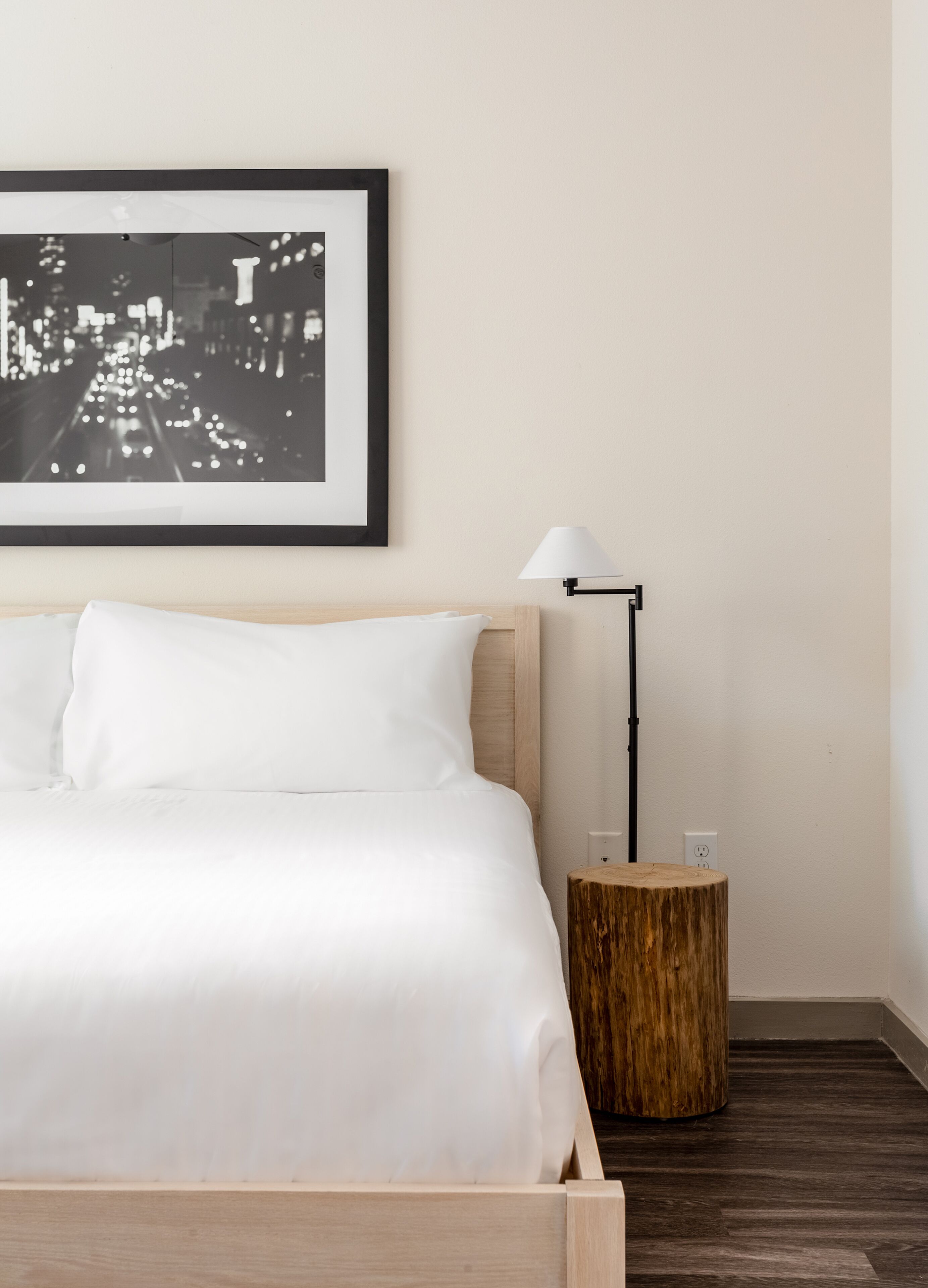 Una cama bien hecha con sábanas blancas, una fotografía en blanco y negro de un paisaje urbano en la pared, una lámpara de pie moderna y un taburete de madera con una taza encima.