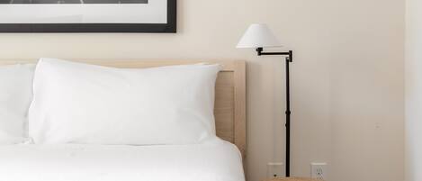 Una cama bien hecha con sábanas blancas, una fotografía en blanco y negro de un paisaje urbano en la pared, una lámpara de pie moderna y un taburete de madera con una taza encima.