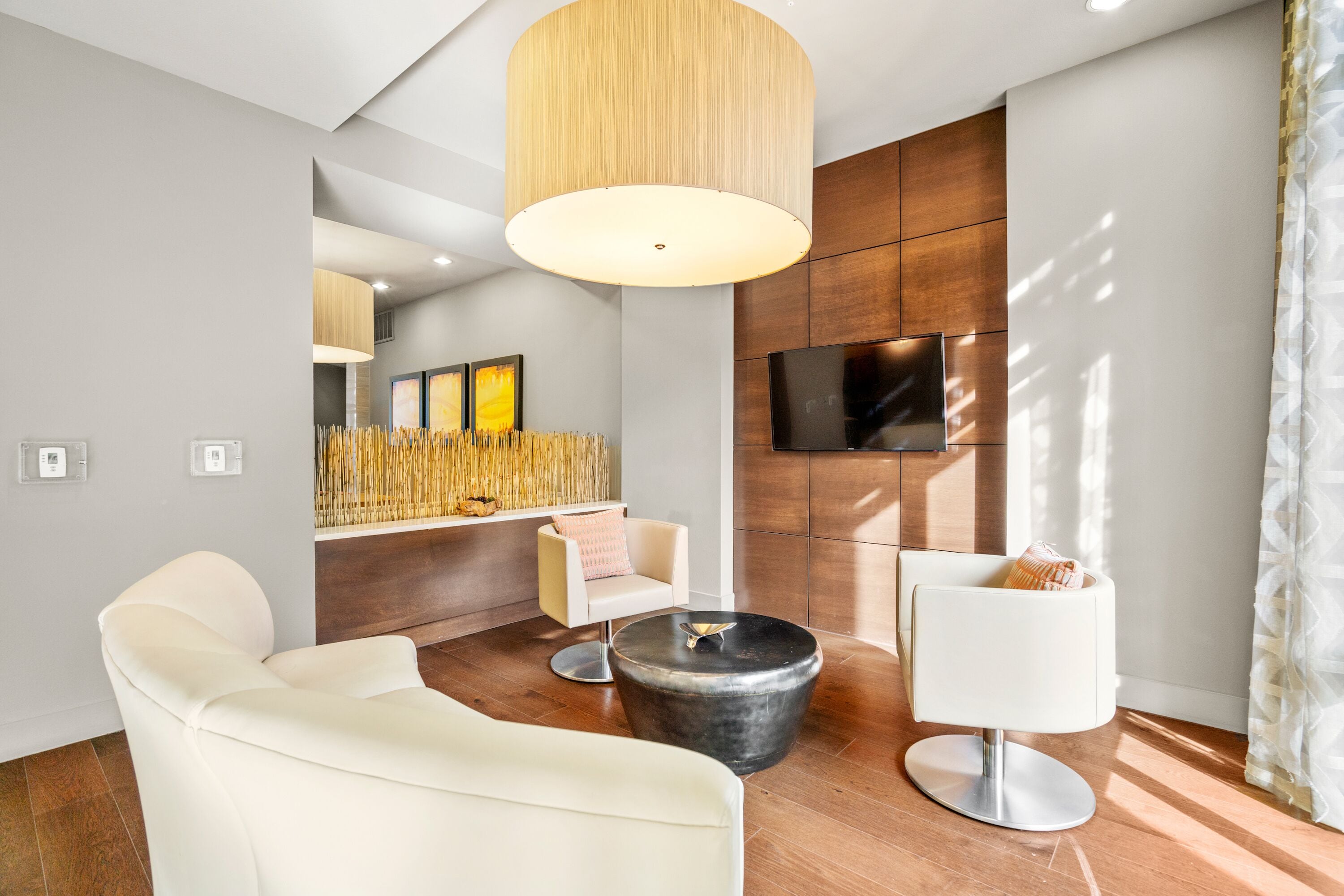Un salon moderne avec une grande table basse ronde, un canapé et un fauteuil. Un téléviseur est fixé au mur et une grande fenêtre est agrémentée de rideaux.