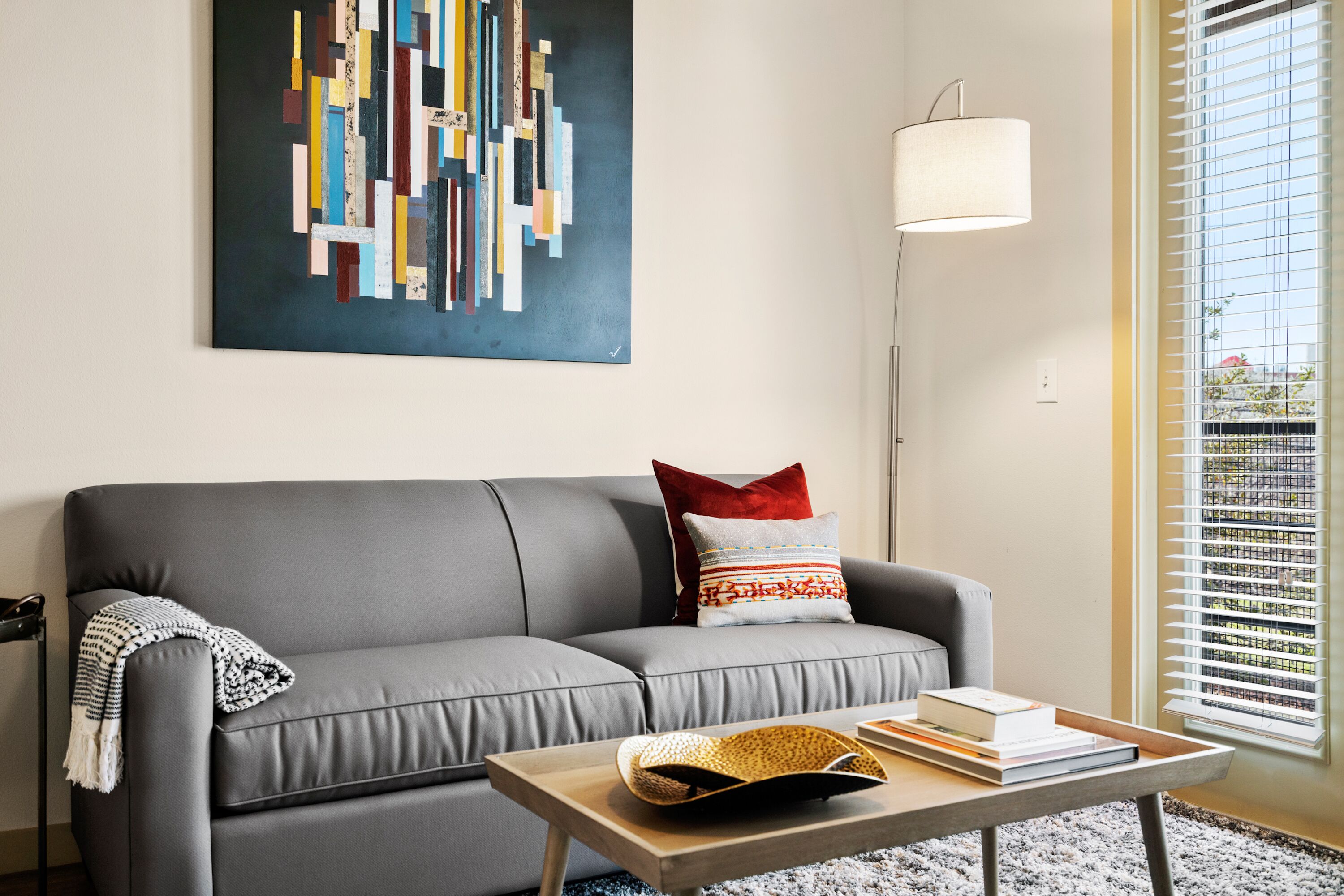 A modern living room with a grey sofa, a coffee table with books and a woven basket, a colorful abstract painting on the wall, and a floor lamp.