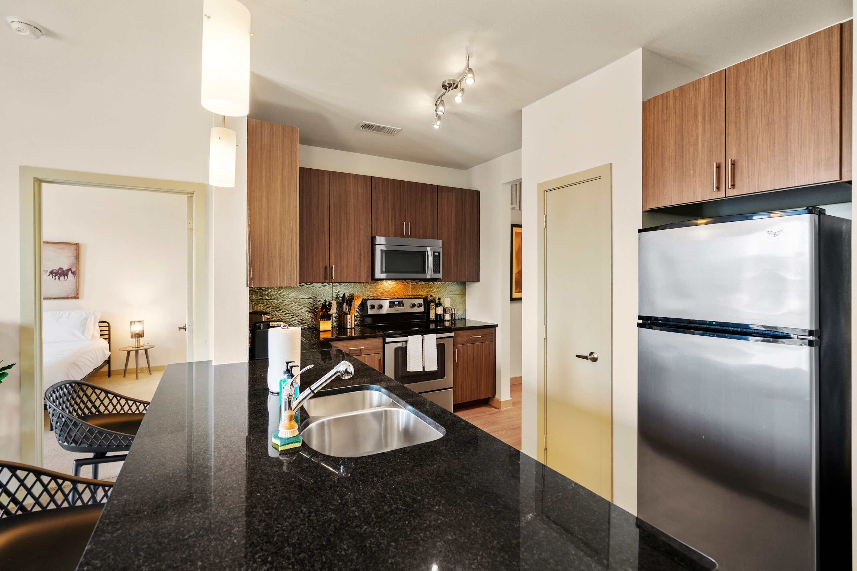 Una cocina moderna con un refrigerador de acero inoxidable, un microondas y un lavamanos. Hay una zona de comedor con una silla negra y una mesita.