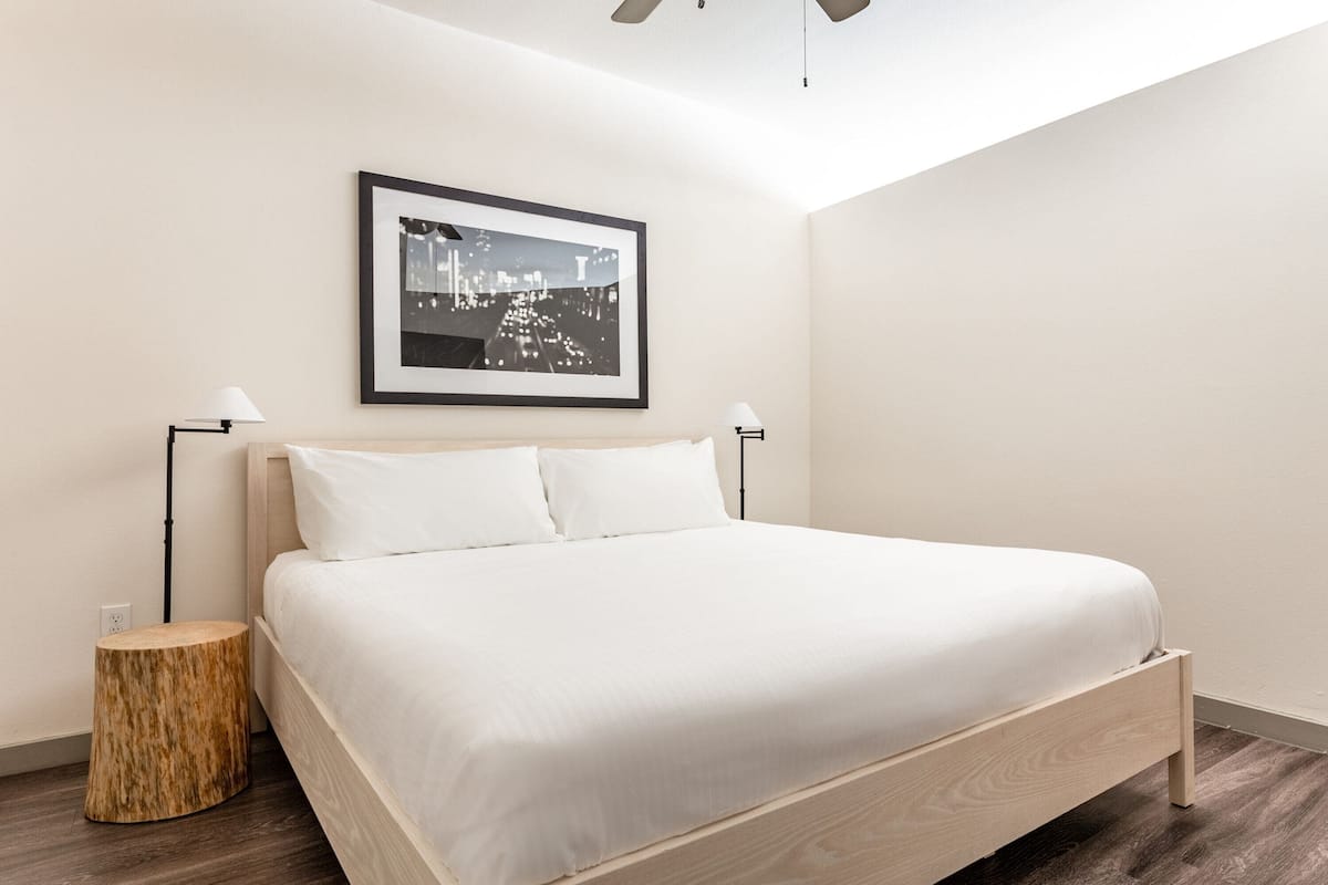 Une chambre à coucher moderne avec un lit, deux lampes de chevet, un tabouret rond en bois et un tableau encadré accroché au mur.