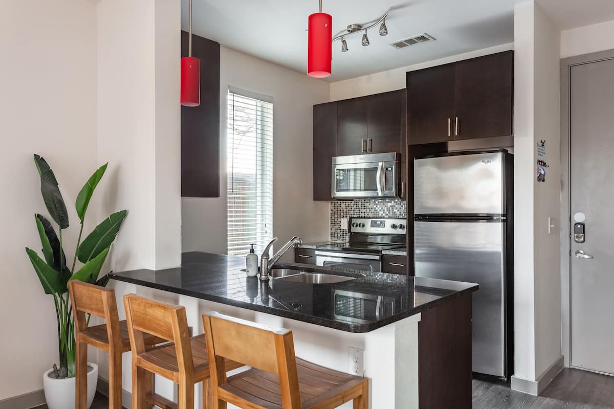 Une cuisine moderne avec des meubles de couleur sombre, des appareils en acier inoxydable, un plan de travail noir et un luminaire suspendu rouge.