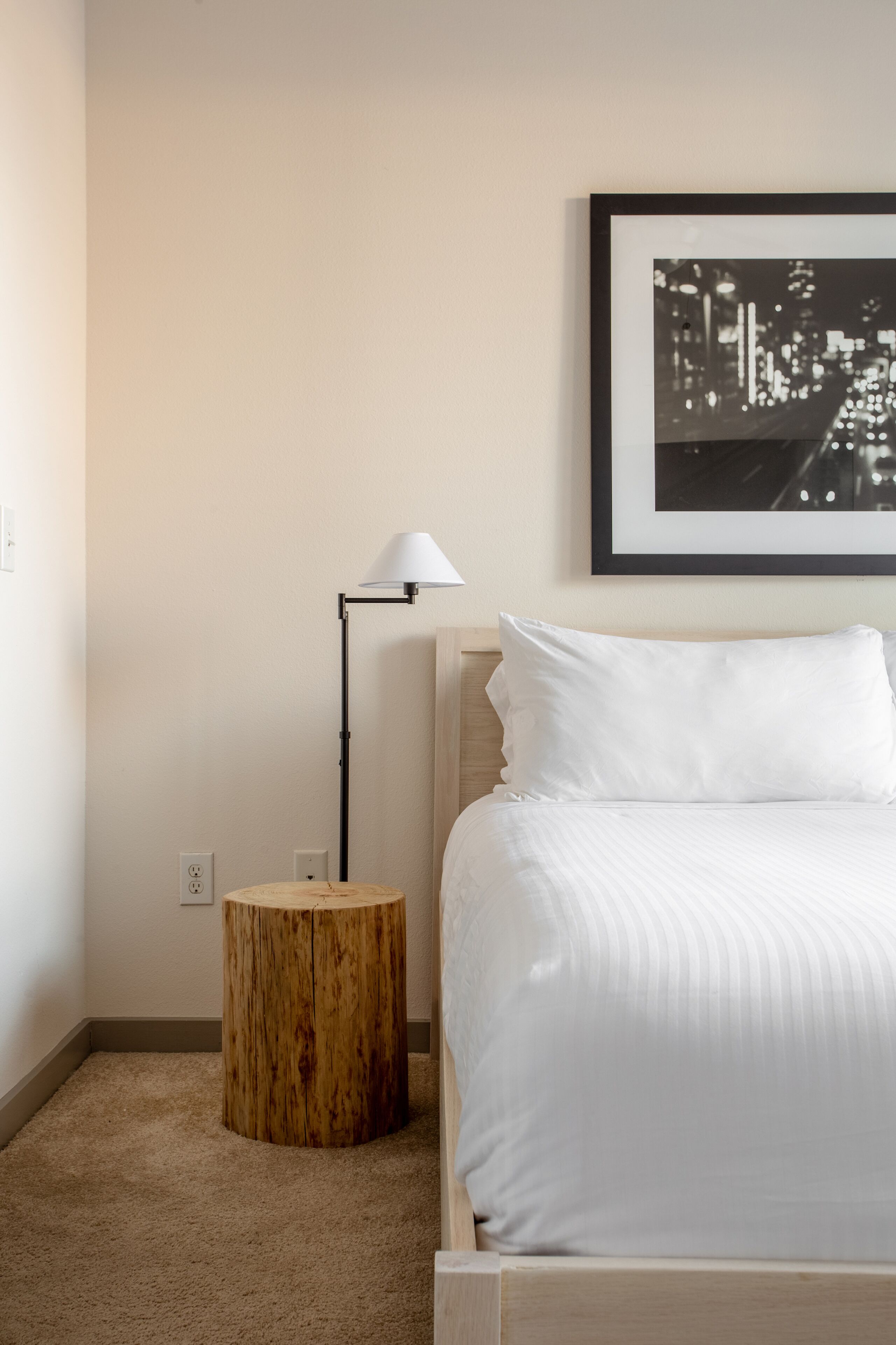 Une chambre d’hôtel bien rangée, avec un lit, une petite table ronde en bois, une lampe sur pied et un tableau représentant une vue de la ville, accroché au mur.