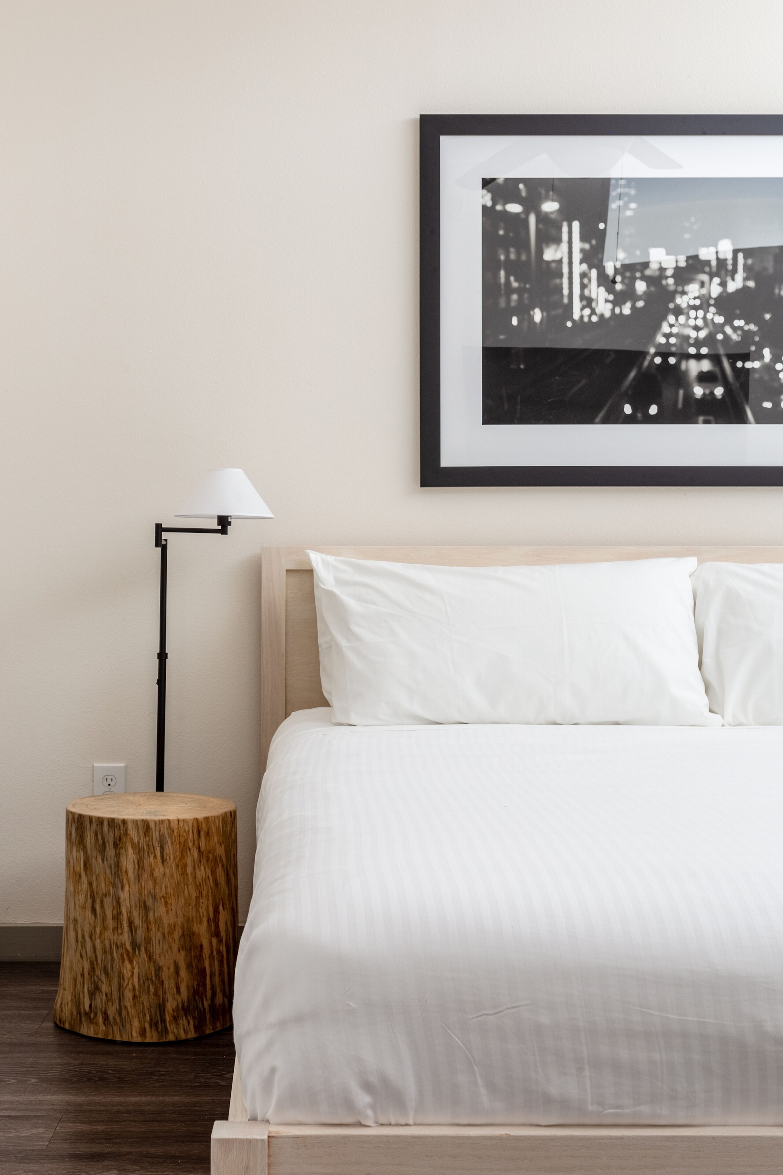 Camera d'albergo ordinata con un letto, un tavolino rotondo in legno, una lampada da terra e un quadro con una veduta cittadina appeso al muro.