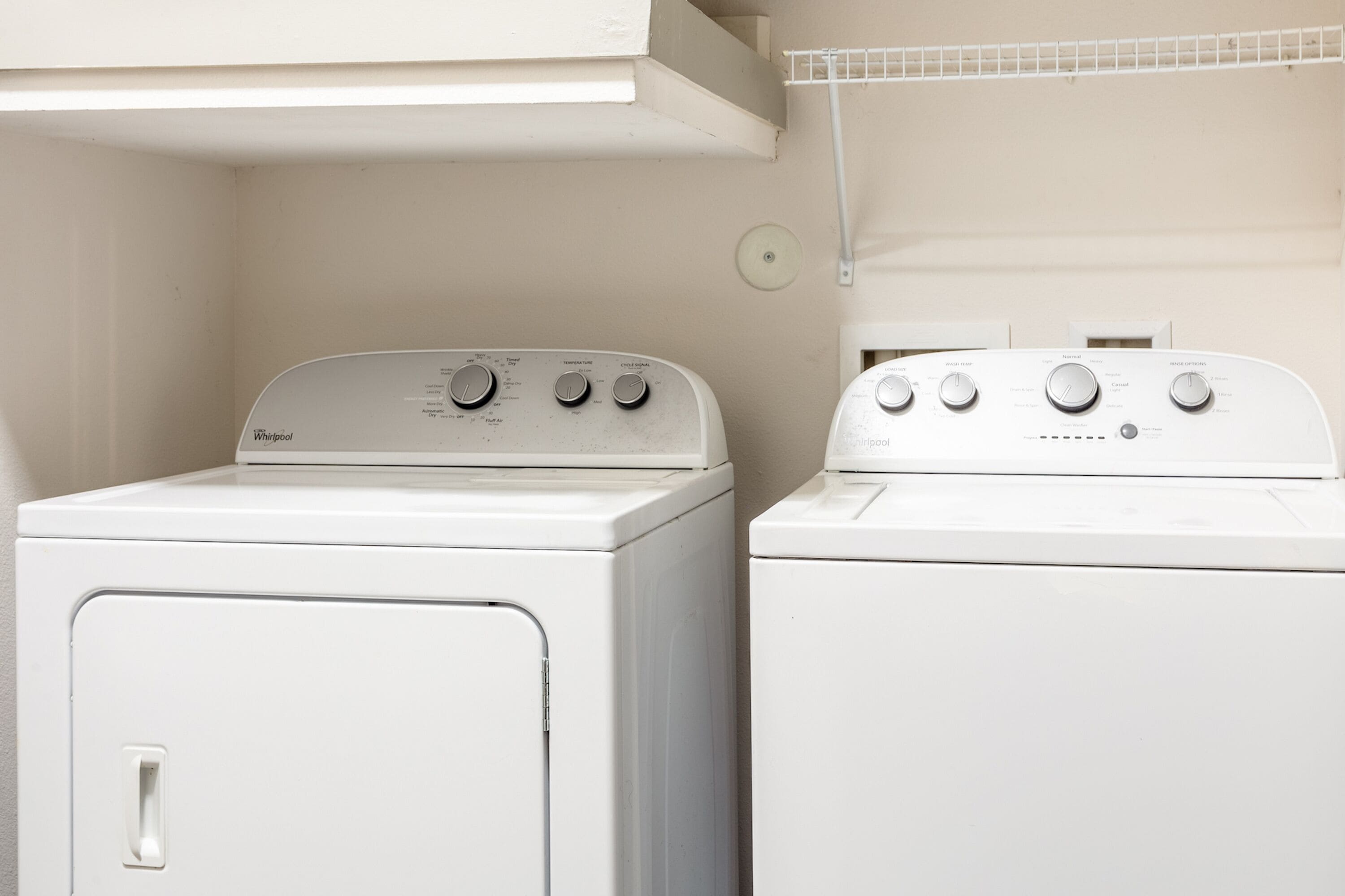 Two white Whirlpool dryers side by side in a laundry room with a white wall and a shelf above.