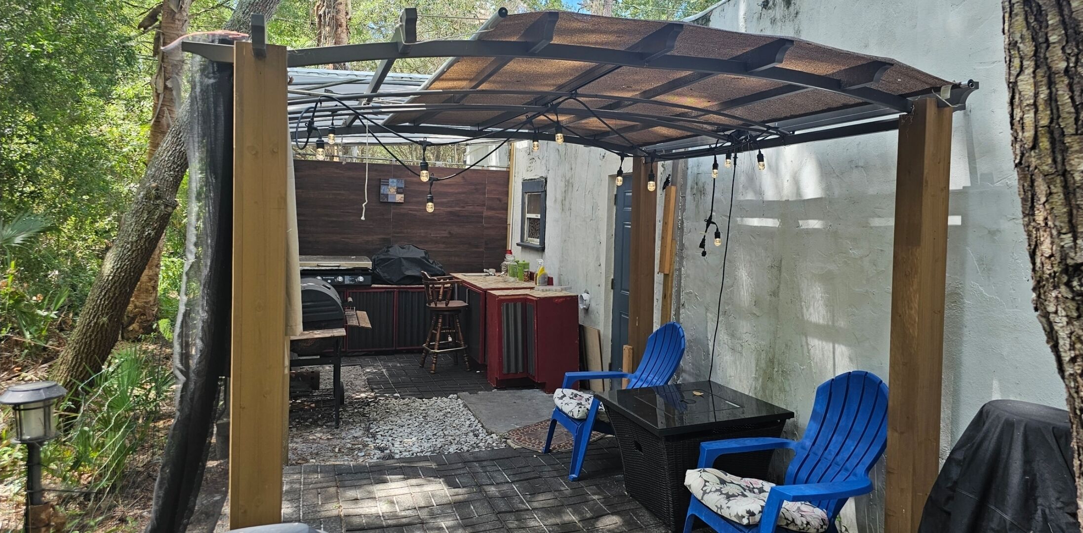 Eine überdachte Terrasse im Freien mit einem Grill, blauen Stühlen und einem Tisch.
